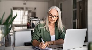 Woman writing on paper sitting at a desk in front of a laptop computer