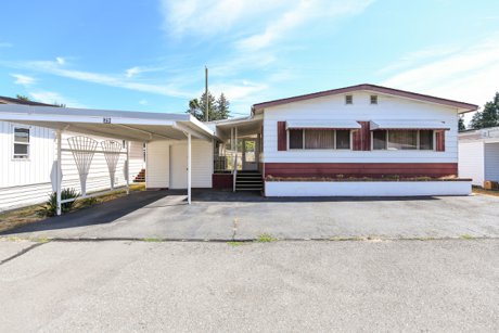 A bright and spacious double-wide manufactured home in the 55+ Little River community, featuring a well-maintained exterior, updated covered porch, mature landscaping, and a peaceful setting surrounded by greenery.